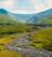 Lush green mountains, against a blue sky, with a babbling brook in the foreground.