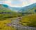 Lush green mountains, against a blue sky, with a babbling brook in the foreground.